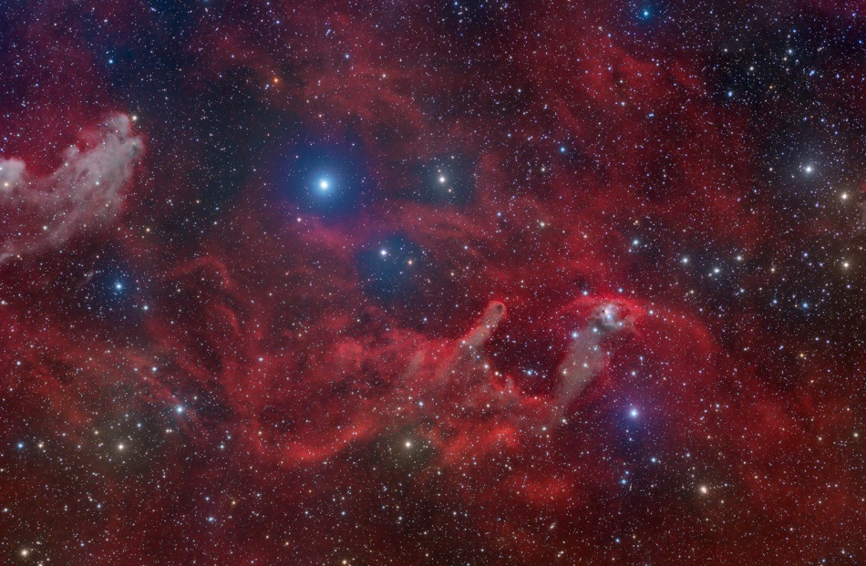 Rote Nebel im Sternbild Orion bilden einen Kontrast zu hellen Sternen, die von einem blauen Schimmer umgeben sind. Die bläuliche Wolke, die links ins Bild ragt, ist das Kinn des Hexenkopfnebels.