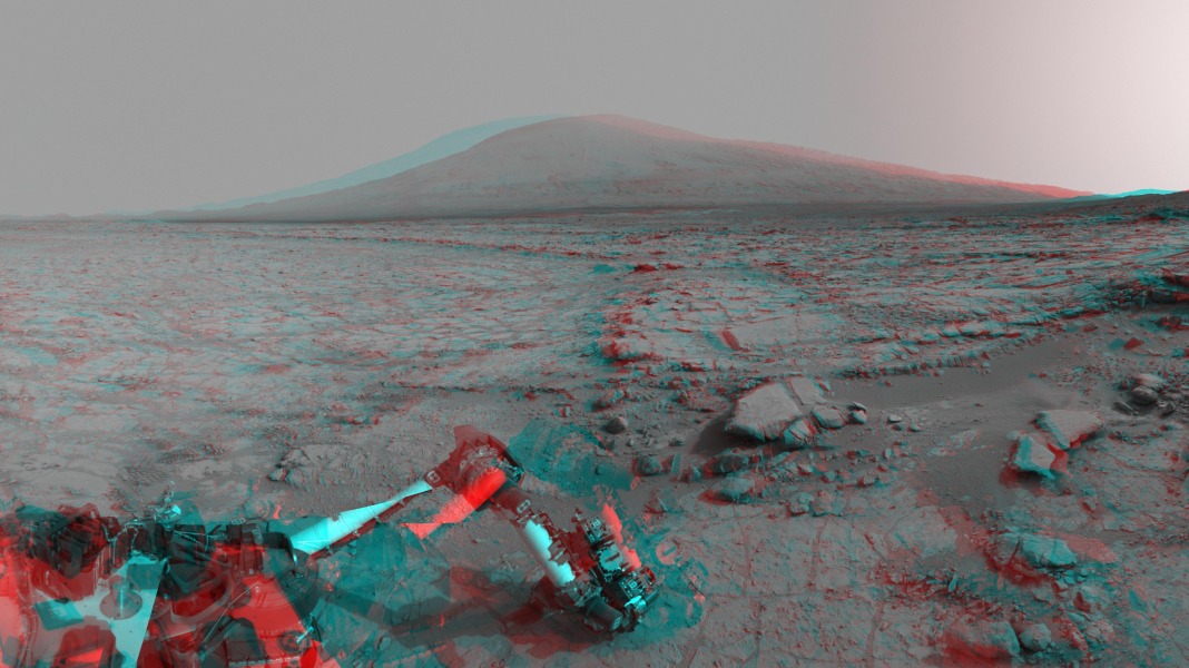 Das Bild ist rot und cyan gefärbt. Mit rot-blauen Brillen wirkt es dreidimensional. Hinten am Horizont ragt Mount Sharp im Krater Gale auf dem Mars auf. Vorne sind Teile des Rovers Curiosity zu sehen.
