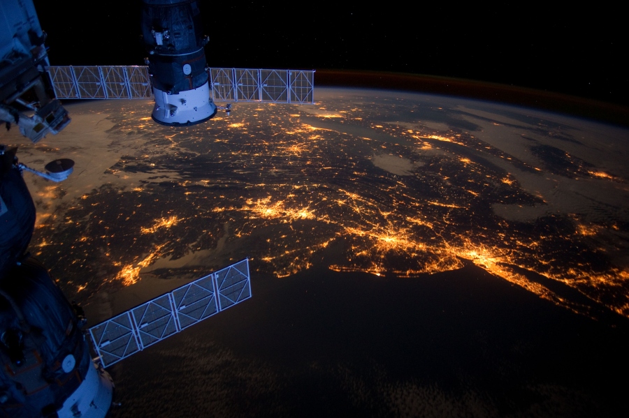 Hinter Solarpaneelen und einer Sojuskapsel an der Internationalen Raumstation ISS leuchten die Lichter der Städte New York, Philadelphia, Baltimore und Washington DC.