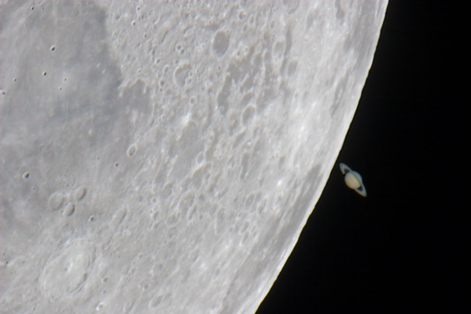 Links ist ein Teil des Mondes riesig im Bild, er füllt einen Großteil des Bildes aus. Rechts am Rand ist gerade der Planet Saturn mit seinen Ringen, der im Vergleich dazu winzig ist, hinter dem Mondrand aufgetaucht.
