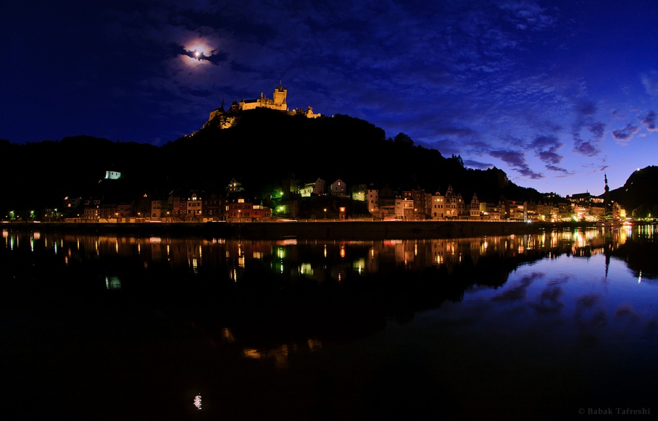Über der Mosel ragt hinter Cochem ein Berg auf, oben steht die historische Burg Cochem aus dem 11. Jahrhundert. Am Himmel zieht eine Wolke über eine Mondkorona. So entsteht der Eindruck einer Fledermaus, die an den Himmel projiziert wurde.