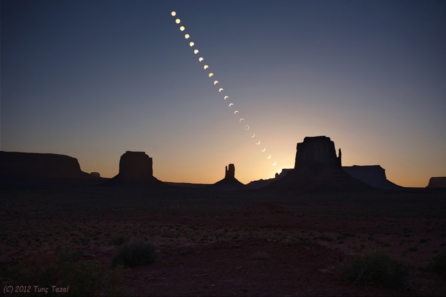 Hinter der markanten Silhouette des Monument Valley mit den berühmten Tafelbergen, den Mittens, sinkt am glasklaren Himmel die Sonne zum Horizont. Dabei wird sie teilweise vom Mond verdeckt.