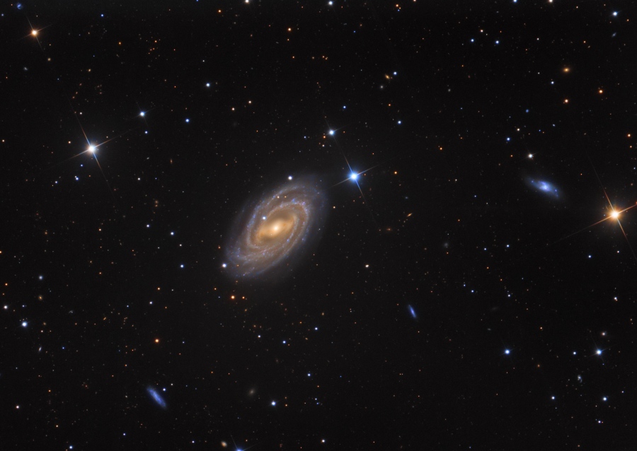 Mitten im Bild liegt schräg eine Spiralgalaxie mit einem markanten Zentralbalken. Auch einige kleine bläuliche Galaxien sind im Bild. Links, in der Mitte und rechts sind drei etwa gleich helle Sterne mit Zacken in einer Linie angeordnet.