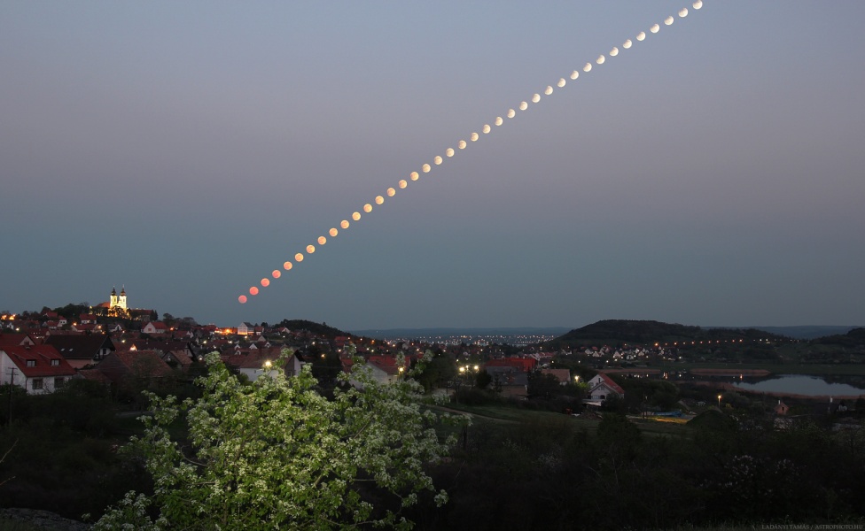 Über einer dämmrigen Landschaft geht der Mond auf. Der Vollmond ist unten rötlich vom Abendrot, weiter oben taucht er ein kleines Stück in den Erdschatten ein. Die Szene zeigt Tihany am Ufer des Balaton.