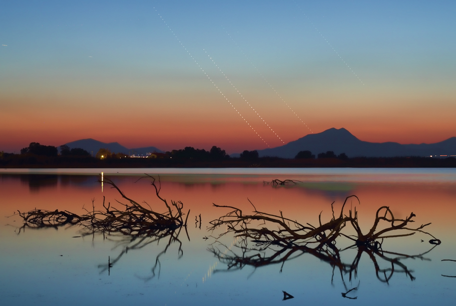 Über dem Salzsee Alikes auf der griechischen Insel Kos ziehen drei Planeten und ein Stern ihre gepunkteten Linien in der Dämmerung. Im Wasser schwimmen Äste oder Wurzeln, hinter dem See ist ein Wald und dahinter ein Berg.