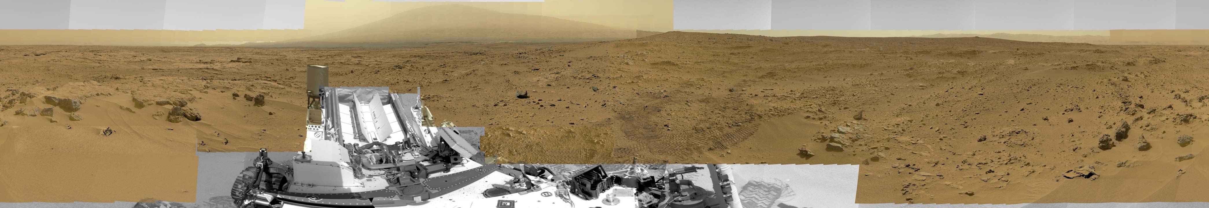 Vorne im breiten Marspanorama breitet sich der Rover Curiosity aus, dahinter am Horizont ragt der Mount Sharp auf.