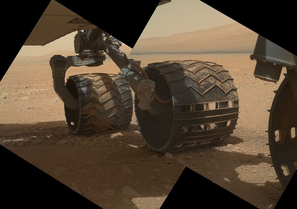 Das Bild zeigt den Blick unter den Marsrover Curiosity. Prominent im Bild sind zwei Räder des Rovers, ein drittes Rad ragt von rechts ins Bild.