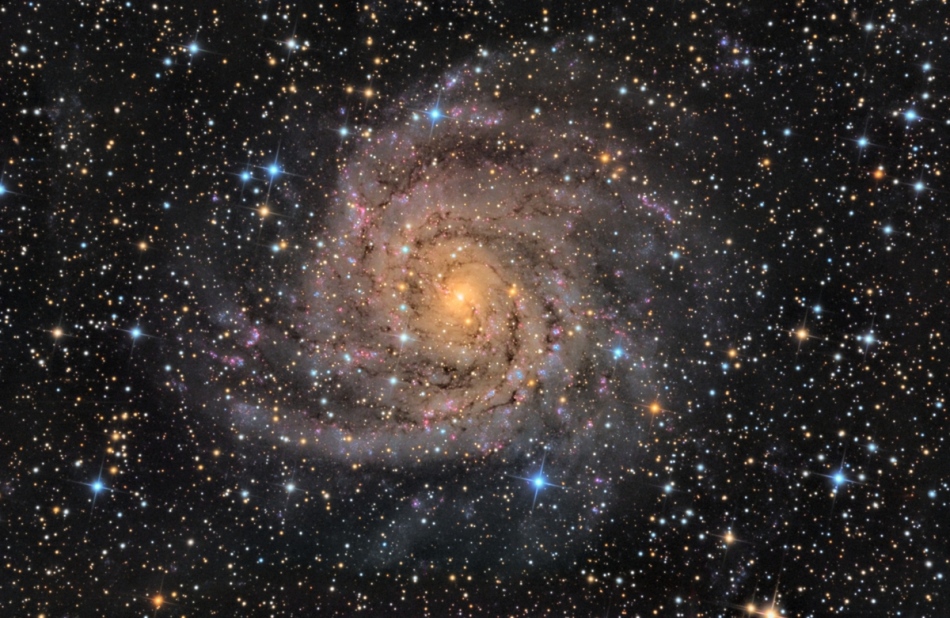 Hinter einem Perlenvorhang aus bunten Sternen leuchtet eine von oben sichtbare Spiralgalaxie, im Bild leuchtet sie gelb mit einigen rötlichen Sternbildungsregionen.