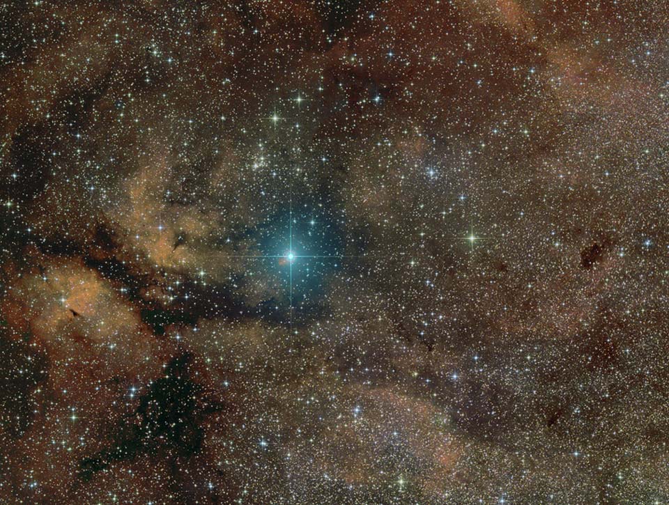 Das Bild ist voller bräunlicher Nebel, Dunkelnebel und dicht gedrängter Sterne. In der Mitte leuchtet ein blauer Stern mit Lichtkreuz.