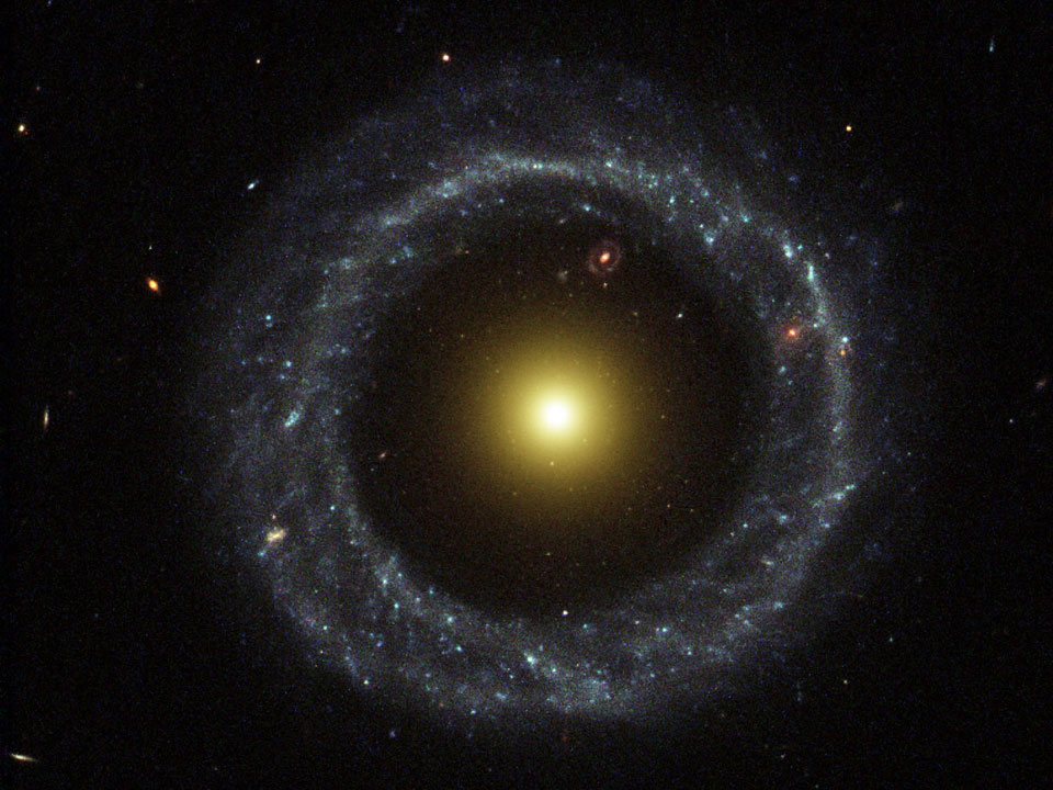Hoags Objekt im Bild ist eine Galaxie, sie hat einen Kranz aus jungen blauen Sternen und eine diffuse, gelb leuchtende Kugel im Inneren. Dazwischen ist eine Lücke, durch die eine weitere Ringgalaxie zu sehen ist.