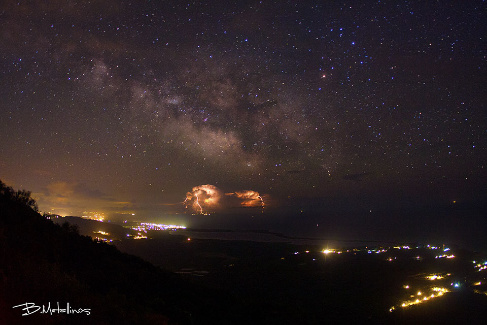 Am Himmel zucken Blitze aus der Milchstraße. Im Vordergrund beleuchten Lichter von Städten um den Korission-See auf der griechischen Insel Korfu die Nacht.
