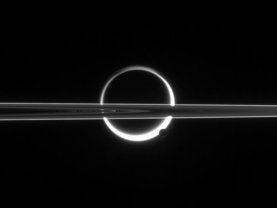 Das seltsam wirkende Bild zeigt den Saturnmond Titan von einem hellen Kranz umgeben. Es ist Gas aus seiner Atmosphäre, das von hinten beleuchtet wird. Unten ragt Enceladus in den Ring. Davor schneiden die schmalen Saturnringe waagrecht ins Bild.