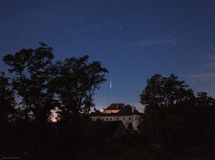 Über dem mittelalterlichen Schloss Albrechtsberg an der Pielach hinter den Silhouetten von Bäumen zischt ein Meteor über den nächtlichen Himmel.
