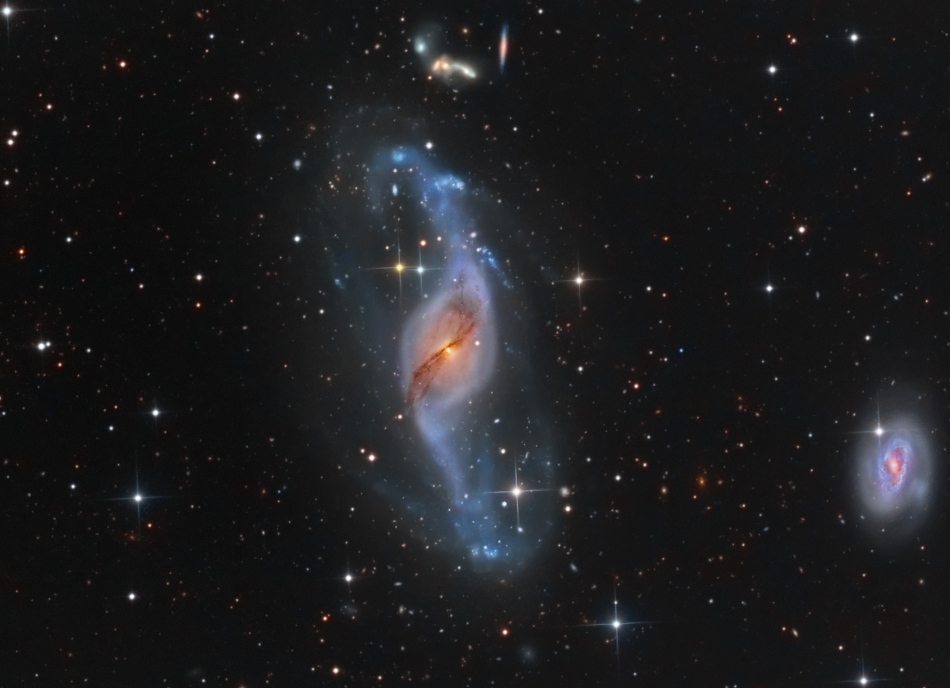 Im Bild sind verzerrte Galaxien im Sternbild Ursa Major verteilt. In der Mitte ist eine senkrecht in die Länge gezogene, verzerrte Spiralgalaxie mit bläulichen Spiralarmen und einem gelblichen Kern, vor dem eine dunkle Staubwolke verläuft.