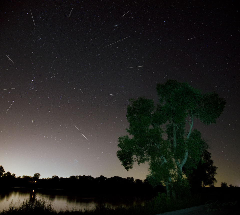 Vorne links ist ein spiegelglattes Wasser, rechts steht ein Baum. Am Himmel steht das Sternbild Perseus, aus dem scheinbar viele Meteore strömen. Auch die Sternbilder Dreieck und Andromeda sind erkennbar.