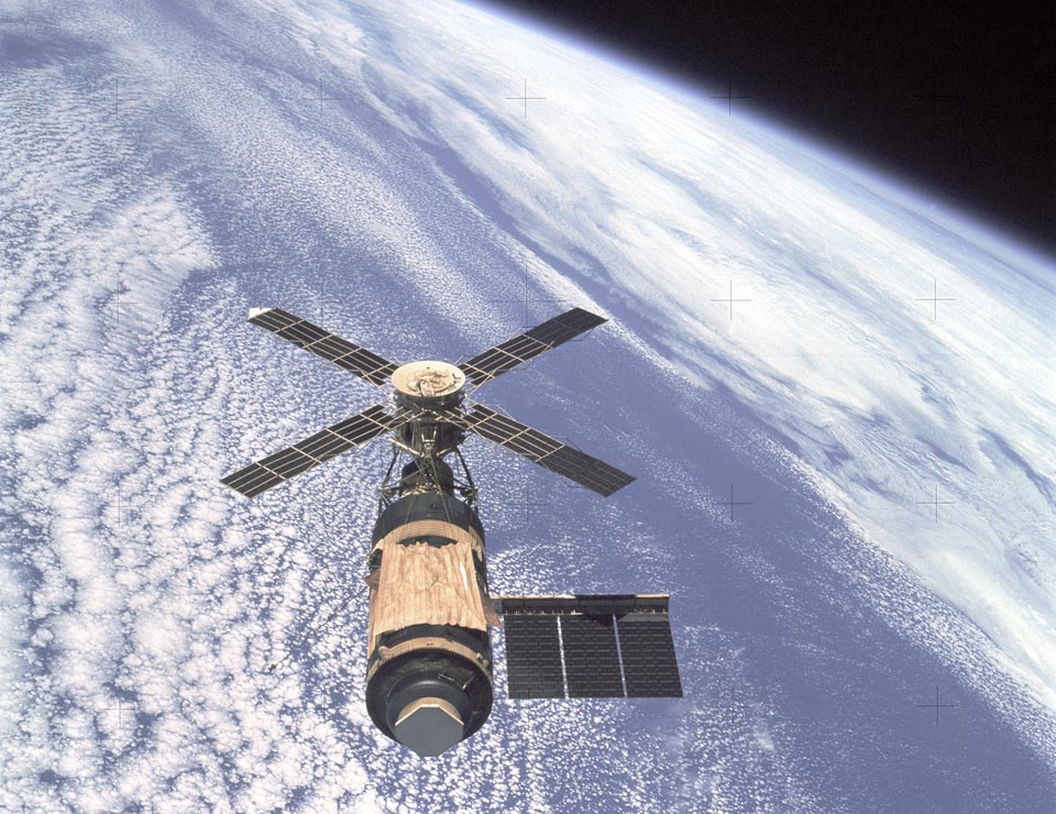 Über der blauen, wolkigen Erde schwebt die Raumstation Skylab. Die Solarpaneele erinnern an die Flügel einer Windmühle.