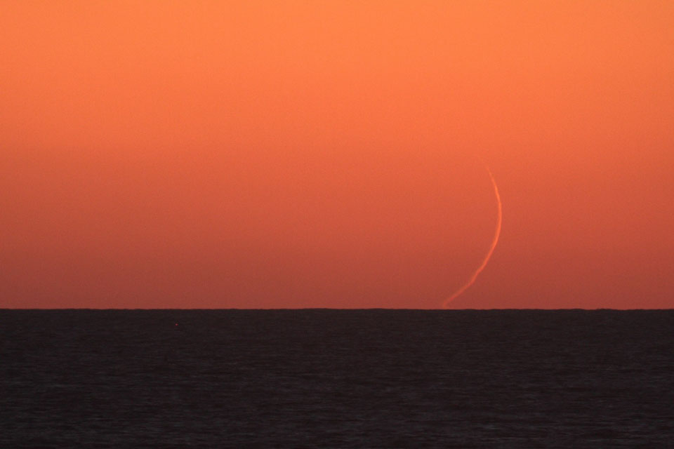 Über den dunklen Horizont des Meeres ragt eine extrem schmale Mondsichel über den Horizont, sie leuchtet hellorange am dunkelorangefarbenen Himmel.