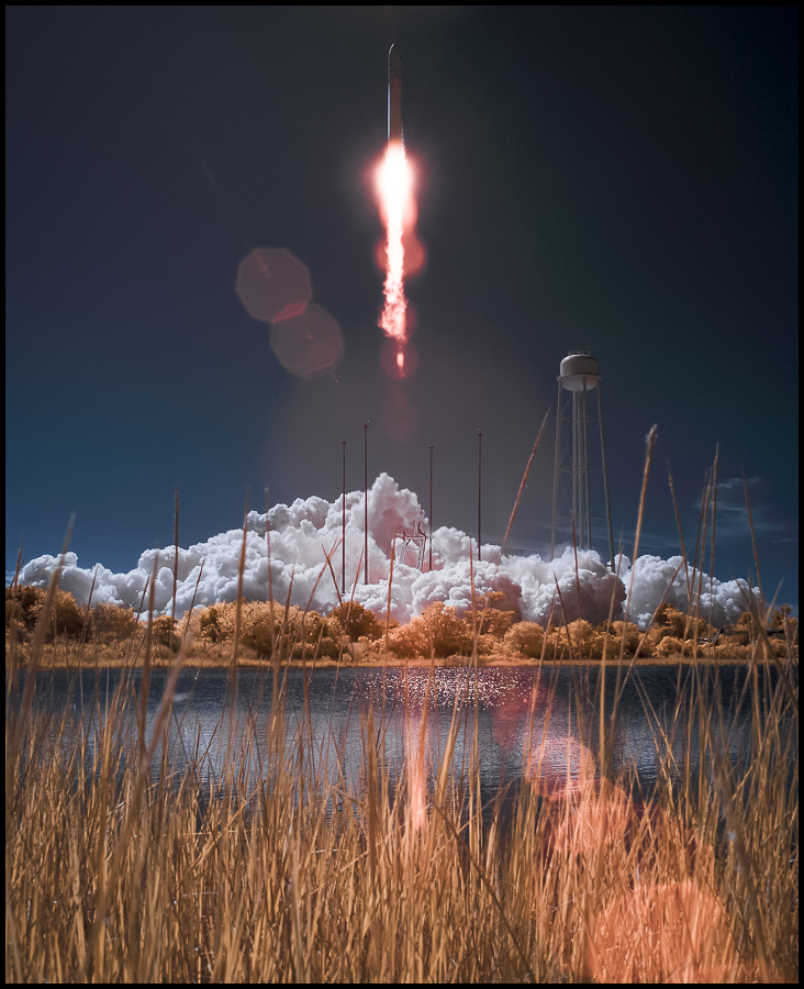 Hinter einem Gewässer, das von Schilf umgeben ist, steigt eine Rakete in den Himmel. Rechts neben dem Feuerschweif steht ein Wasserturm. Durch die Infrarotaufnahme wirkt das Bild eigenartig blass.