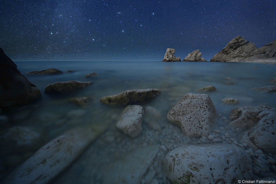 Hinter dem felsigen Ufer am Meer ist über dem Horizont ein Sternenhimmel zu sehen.