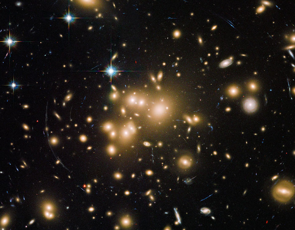 Helle, verschwommen wirkende Galaxien drängen sich in der Mitte dieses Bildes. Im Umkreis sind blaue, schmale Bögen zu finden, es sind Galaxien, deren Bilder von der Gravitation des Haufens verzerrt wurden.