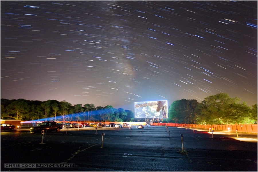 Über einem Autokino leuchten Strichspuren von Sternen und die Milchstraße. Auf der Leinwand läuft ein Film mit der Internationalen Raumstation ISS.