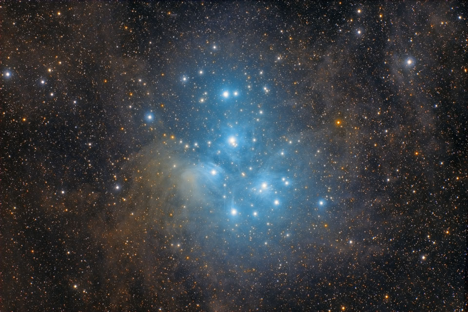 Das Bild ist von Sternen übersät, zwischen denen Nebel verlaufen. In der Mitte drängen sich viele sehr helle Sterne, die von blau beleuchteten Nebeln umgeben sind, es sind die Plejaden, auch M45 genannt.