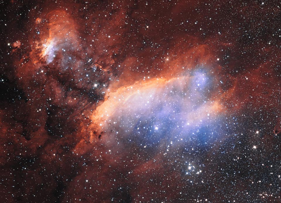 Im Bild leuchtet ein blauer Nebel, der links oben von einem orangefarbenen Grat begrenzt ist. Links oben ist ein kleinerer orangefarbener Nebel. Der Hintergrund ist zart mit Sternen gesprenkelt.