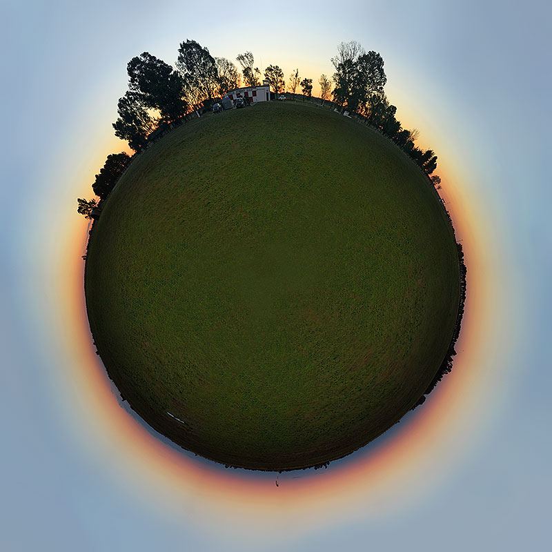 Der kleine Planet im Bild ist von einem orangefarbenen Abendrot umgeben. Am unteren Rand steigt der Erdschatten auf, oben stehen Bäume und in der Mitte ein Haus.