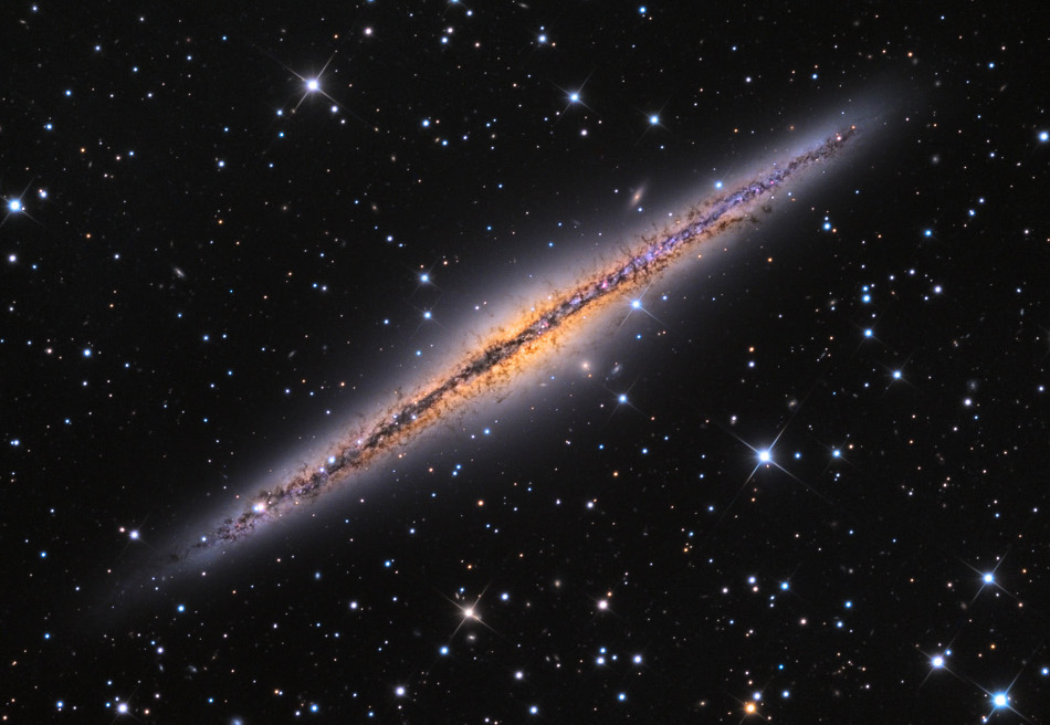 Mitten im Bild verläuft eine spindelförmige Galaxie, die wir von der Kante sehen. Durch die Mitte verläuft eine dunkle Staubbahn. Rundherum sind Sterne verteilt.