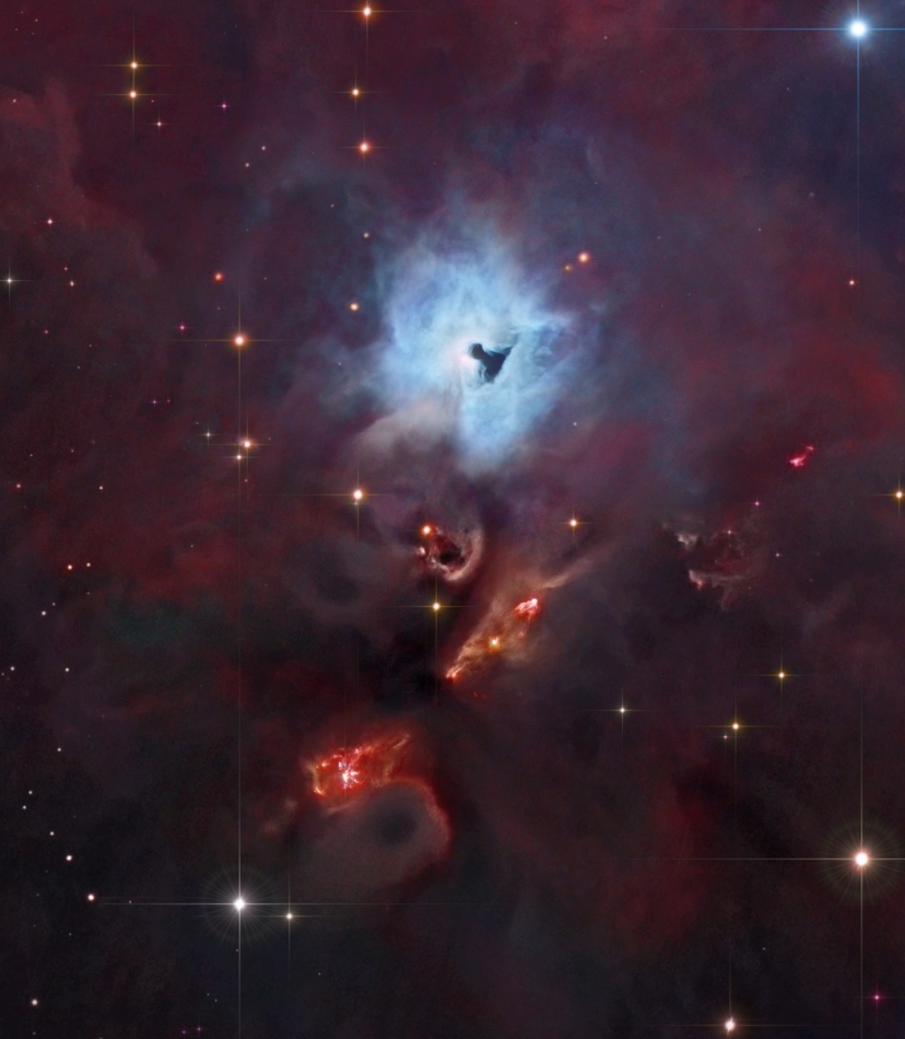 Mitten im Bild leuchtet ein blauer Reflexionsnebel, in den eine schlüssellochförmige dunkle Form wie ein seitlich geneigtes T gekerbt ist. Darunter befinden sich die Bugwellen zweier rot leuchtender Herbig-Haro-Objekte.