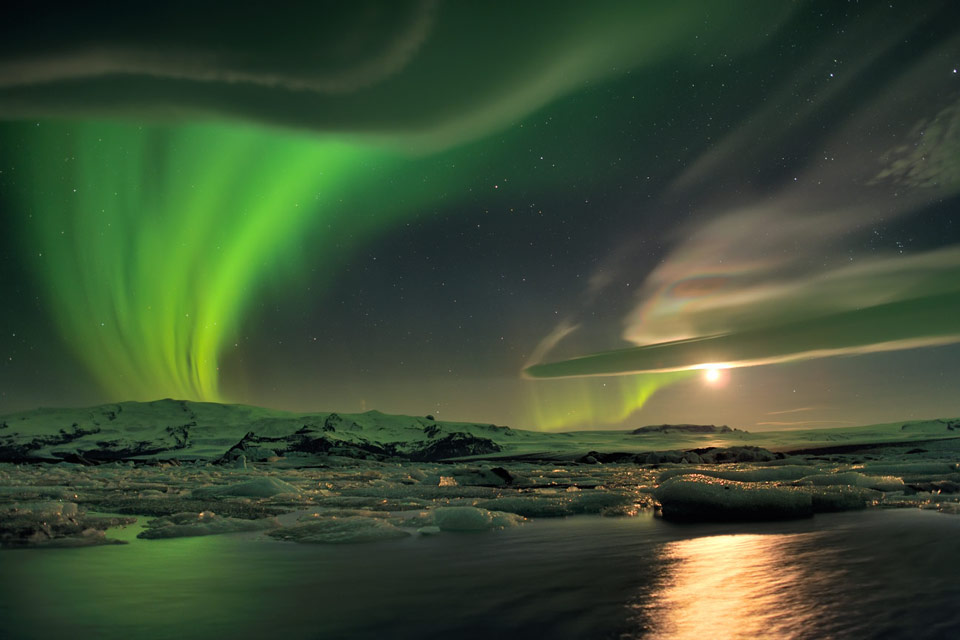 Hinter einem Gletscher und einem See leuchten grüne Polarlichter und der Mond.