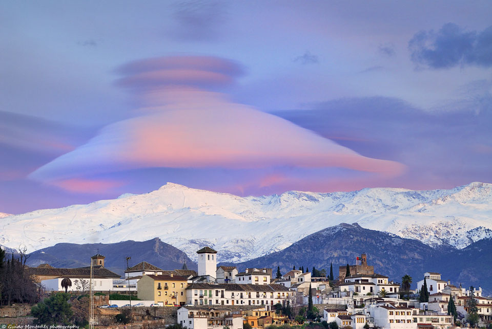 Über einem verschneiten Gebirge ragt eine riesige glockenförmige Wolke auf. Vorne ist eine Stadt im spanischen Granada zu sehen.