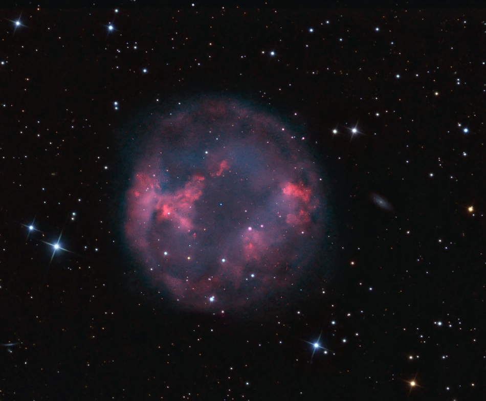 In der Mitte ist ein runder planetarischer Nebel. Einige rot leuchtende Stellen wurden mit Schmalbandfiltern betont. Rechts hinter dem Nebel ist eine kleine Galaxie.