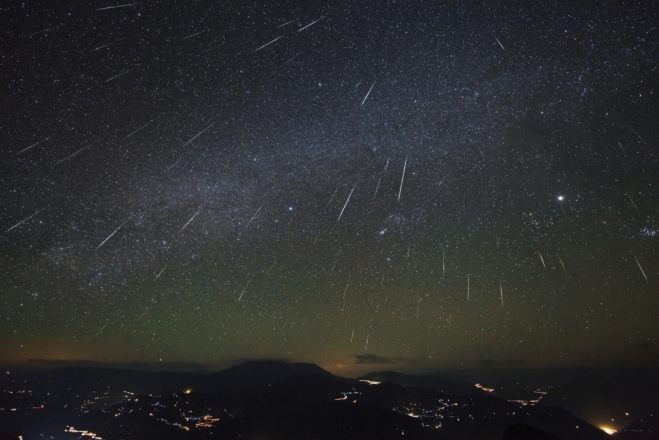 Über einer halbwegs dunklen Landschaft fallen Meteore vom Himmel. Sie strömen von einem Punkt aus, der über dem oberen Bildrand liegt. Am Himmel sieht man Jupiter, das Sternbild Orion und die Milchstraße.