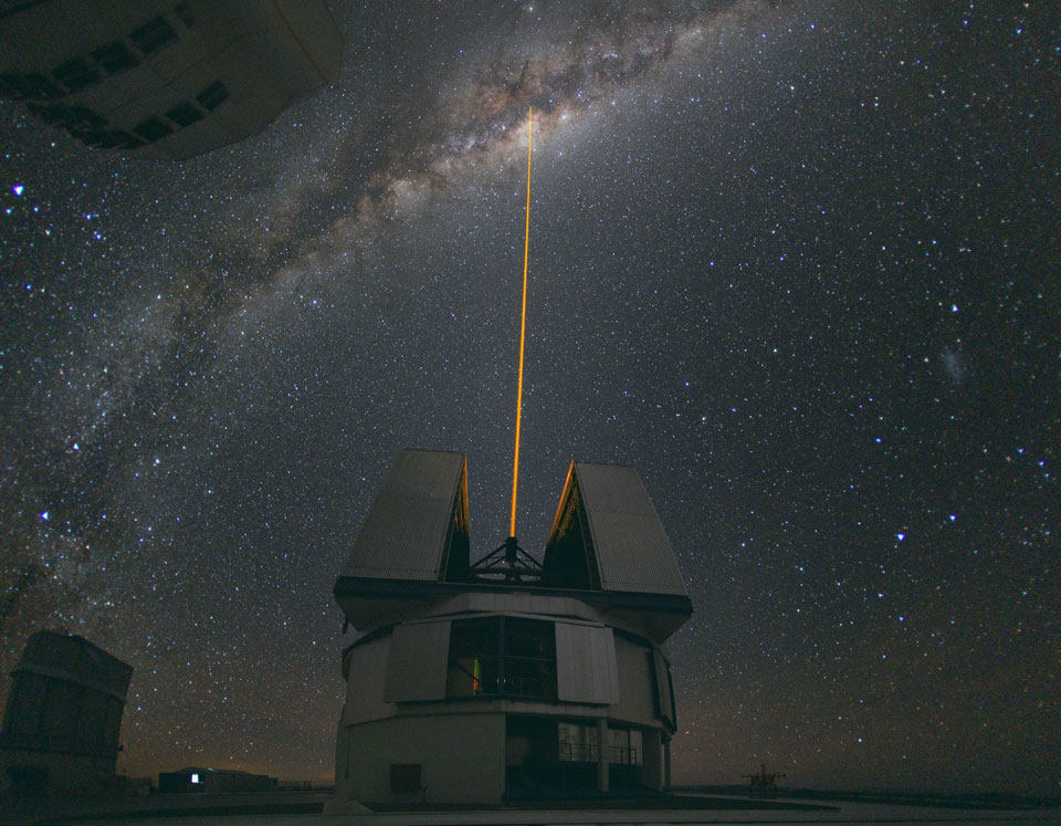 Unten in der Mitte steht ein VLT-Teleskop mit geöffneter Kuppel. Es schießt einen Laserstrahl nach oben zum Zentrum der Milchstraße, das sich oben im Bild befindet. Genaue Erklärung im Text.