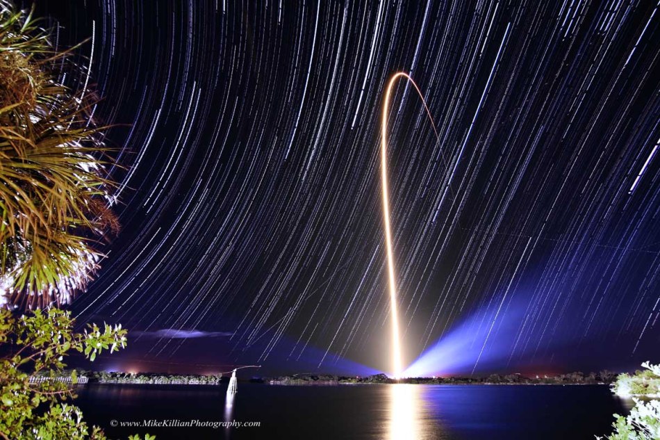 Über einem Wasserbecken verlaufen lange Strichspuren am Himmel. In der Mitte steigt der leuchtende Bogen einer startenden Rakete auf.
