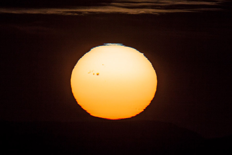 Die Sonne ist von der Lufthülle der Erde stark verzerrt. Links ist eine große Sonnenfleckengruppe, oben ein blauer Rand, unten ein roter.