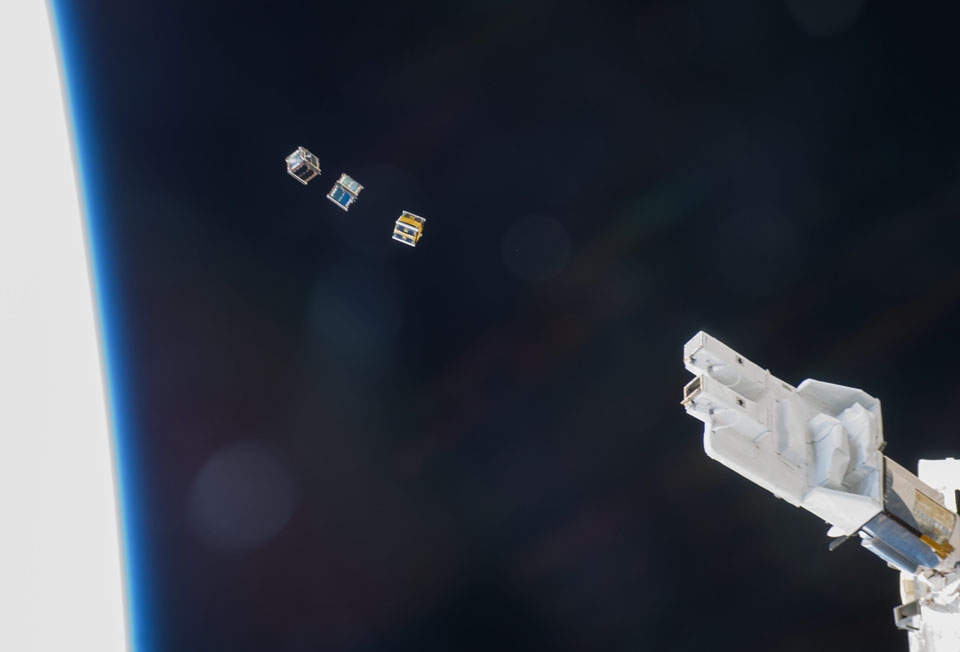 Aus einer Vorrichtung rechts wurden drei würfelförmige Kleinsatelliten ausgestoßen, am rechten Bildrand ist die Silhouette der Erde mit blauem Rand zu sehen.
