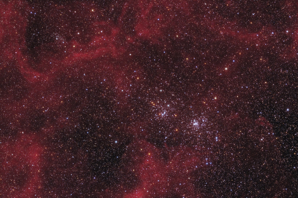 In der Mitte befindet sich der berühmte Doppelsternhaufen h und χ Persei, auch NGC 869 (rechts) und NGC 884. Im Hintergrund sind rote Wasserstoffwolken zwischen dicht gesäten Sternen verteilt.