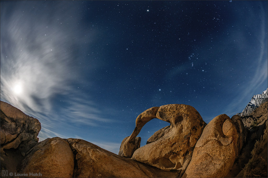 Über dem Möbius Arch in den Alabama Hills östlich vom Mount Whitney in der Sierra Nevada leuchten Sterne und Jupiter am kristallklaren blauen Nachthimmel. Links geht der Mond hinter Wolken auf. Rechts oben steht der Jäger Orion.