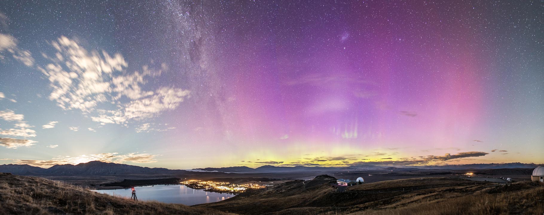 Die Himmelslandschaft über dem neuseeländischen Lake Tekapo hat viel zu biegen: Die Milchstraße, Wolken und Strahlenbüschel, die vom Mond ausgehen, ein Polarlicht und die Große Magellansche Wolke. Links vorne ist der See, rechts stehen Kuppeln des Mount-John-Observatoriums.