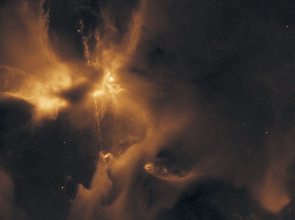 Im Bild sind gelb und orange leuchtende Nebel vor einem dunklen Hintergrund verteilt. Links schießt ein Strahl aus einer hellen Stelle hervor, er bildet ein Herbig-Haro-Objekt.