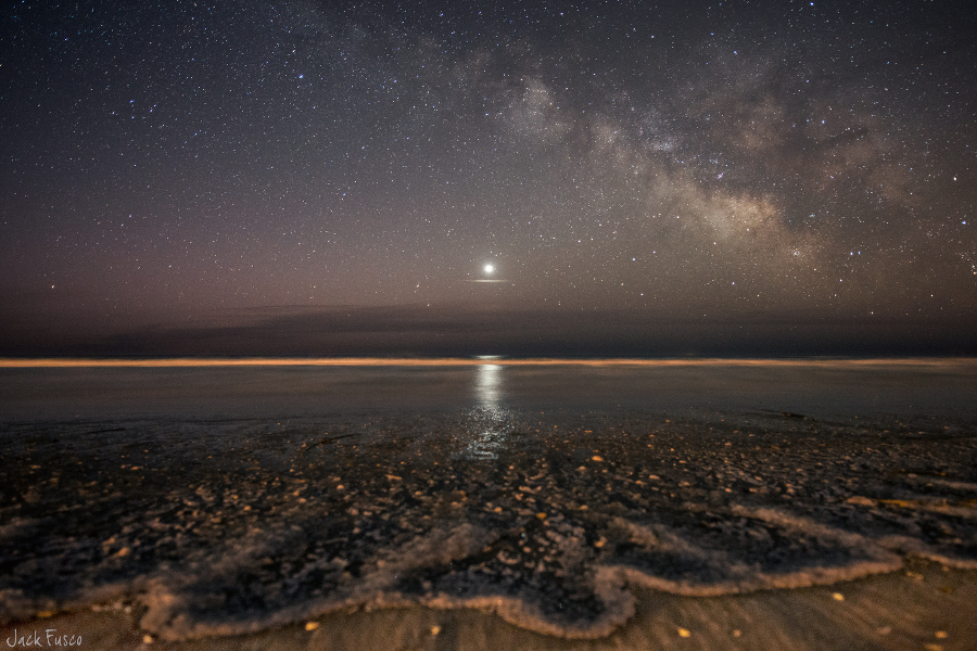 Über dem Horizont strahlt die Venus unter der Milchstraße am sternklaren Himmel. Vorne spült das Meer Wellen an den Sandstrand.