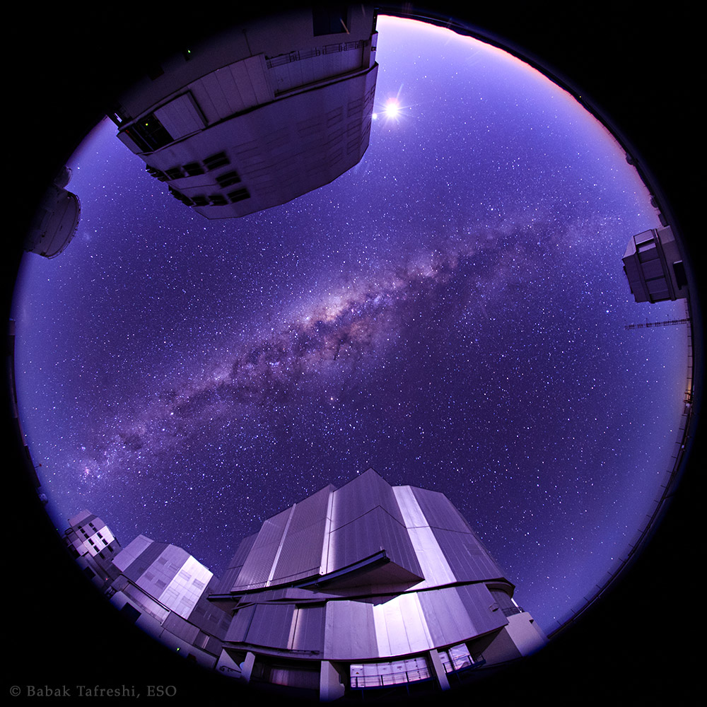 Die Fischaugen-Ansicht zeigt den ganzen dunkelblauen Himmel am Paranal in der Atacama in Chile. In der Mitte verläuft diagonal die Milchstraße. Am Rand stehen die vier 8-Meter-Teleskope des VLT. Oben gleißen Mond und Venus.