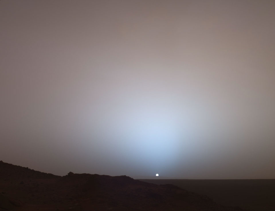 Über dem Horizont geht auf dem Mars eine sehr kleine Sonne unter. Weil die Atmosphäre viel Staub enthält, wird viel blaues Licht nach vorne gestreut.