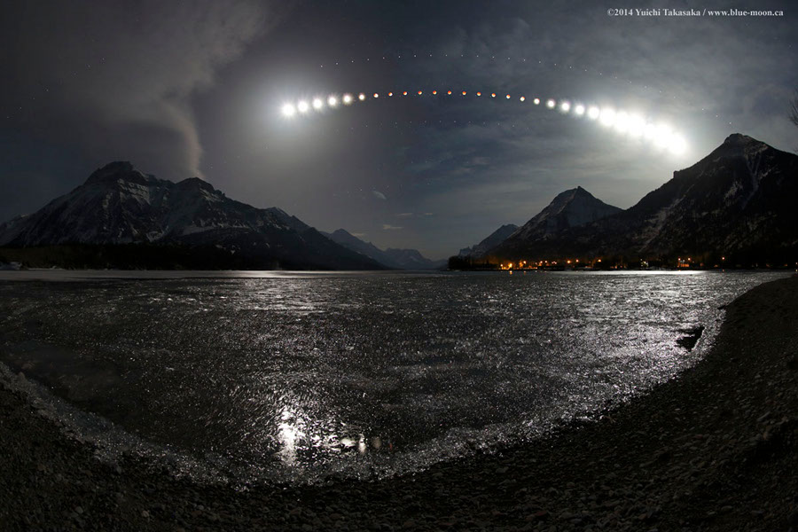 Über einem Gewässer und hinter spitz aufragenden Bergen wandert ein helles Himmelslicht, das in der Mitte dämmrig rot leuchtet. Es ist der Mond im Laufe einer totalen Mondfinsternis.