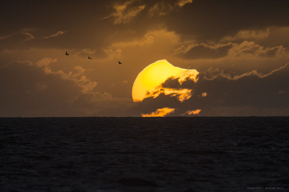 Über dem Meer geht hinter Wolkenfetzen die Sonne unter. Ein kleines Stück ist vom Mond bedeckt, sodass man rechts oben eine Kerbe in der Sonne sieht.