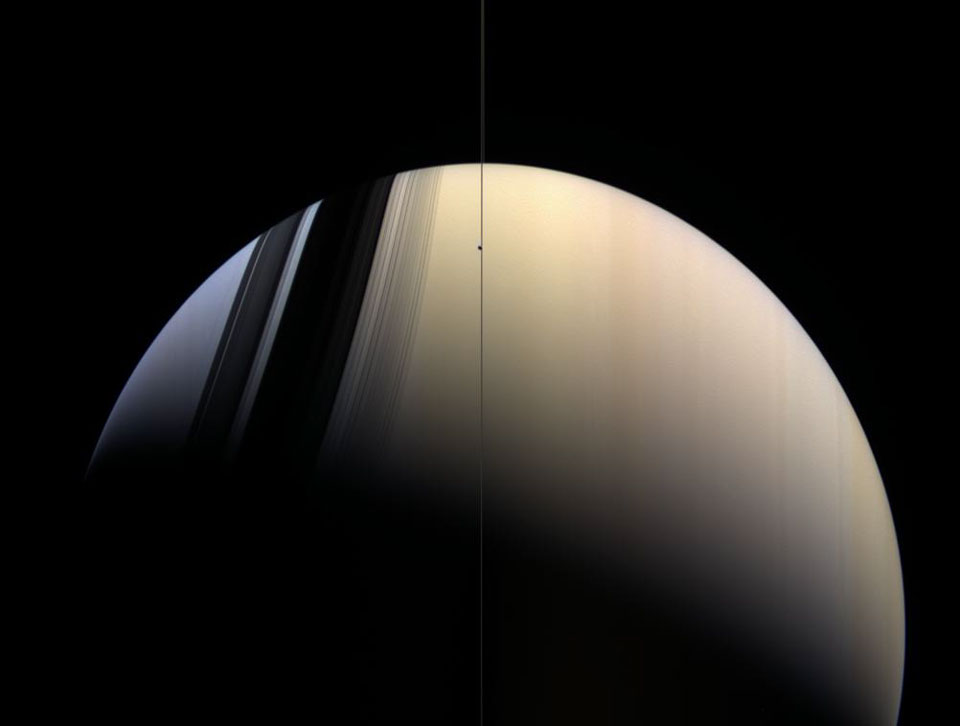 Saturn ist fast halb beleuchtet. Die Ringe verlaufen senkrecht im Bild und sind kaum zu sehen. Sie werfen ihre Schatten nach rechts auf die blaue Nordhalbkugel. Rechts leuchtet Saturn golden.