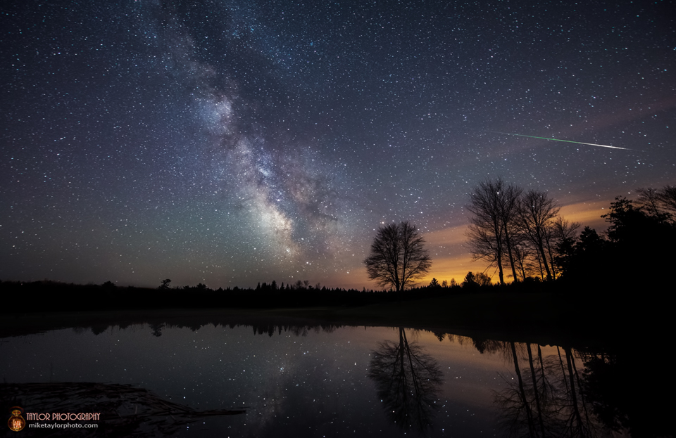 Der Himmel mit der markanten Milchstraße, die in der Mitte aufsteigt, spiegelt sich im Wasser eines ruhigen Teichs. Er ist von einem Wald umgeben, den man nur als dunkle Silhouette sieht. Rechts über einigen Bäumen, die aus dem Wald aufragen, zischt ein Meteor der Eta Aquariiden über den Himmel.