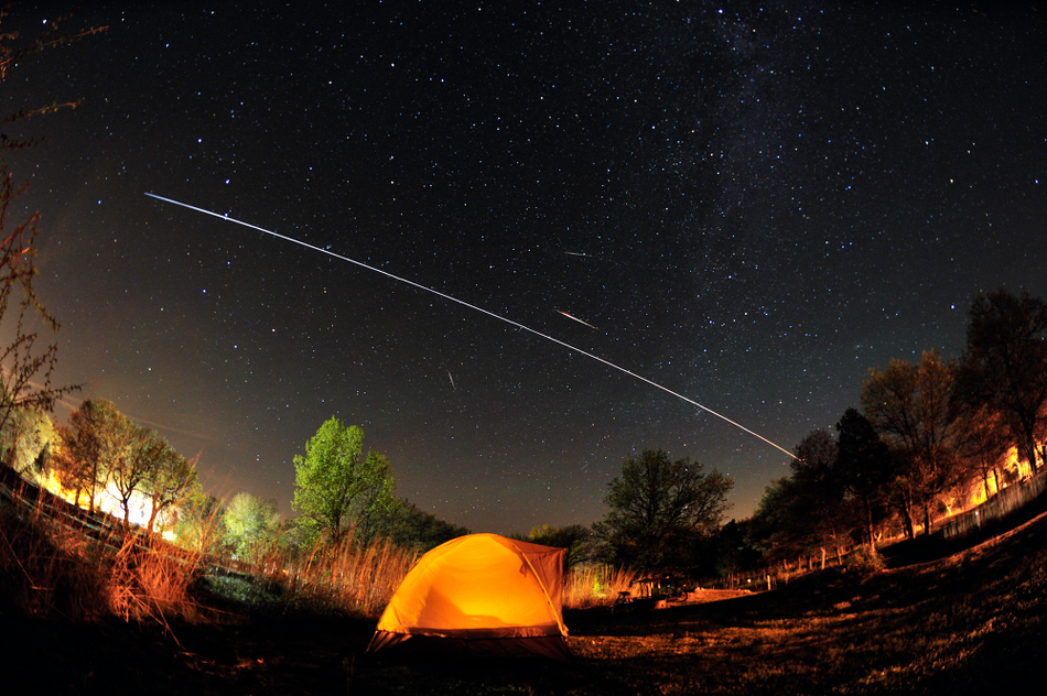Über einem beleuchteten Zeltlager am Eriesee ziehen am sternklaren Himmel leuchtende Spuren. Die kurzen stammen von Meteoren der Camelopardaliden, die lange helle ist die Internationale Raumstation ISS.